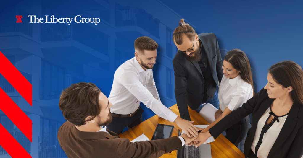 A group of five people huddle around a wooden table, placing their hands together in a gesture of unity; The Liberty Group logo is in the top left corner.