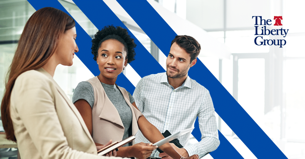 Two women and a man huddle to speak in an office; The Liberty Group logo is in the top right corner.