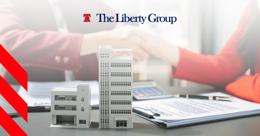 Two people shaking hands over a desk with small building models, documents, and a calculator; The Liberty Group logo is at the top center.