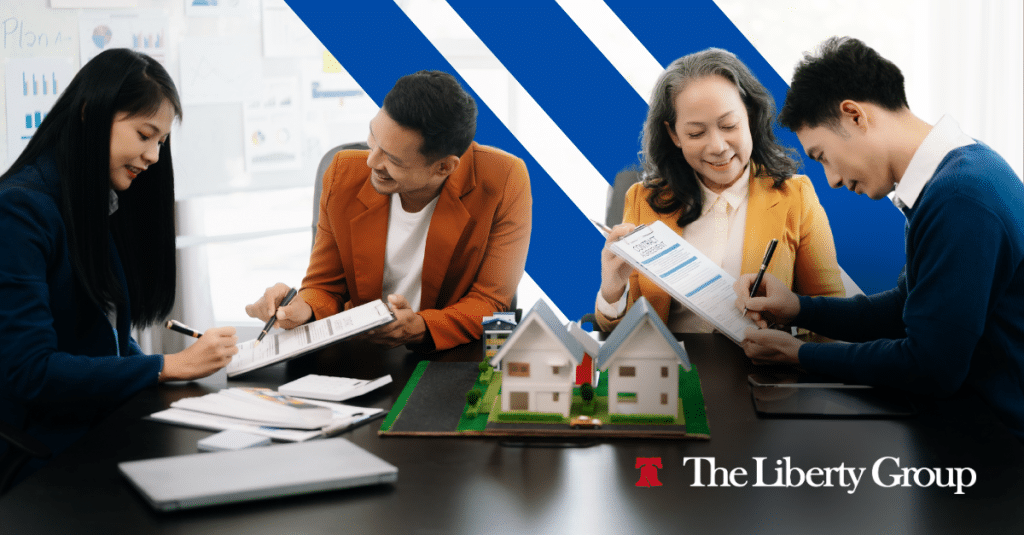 Four people sit at a table reviewing documents, with a small model of houses on the table; The Liberty Group logo is in the bottom right corner.