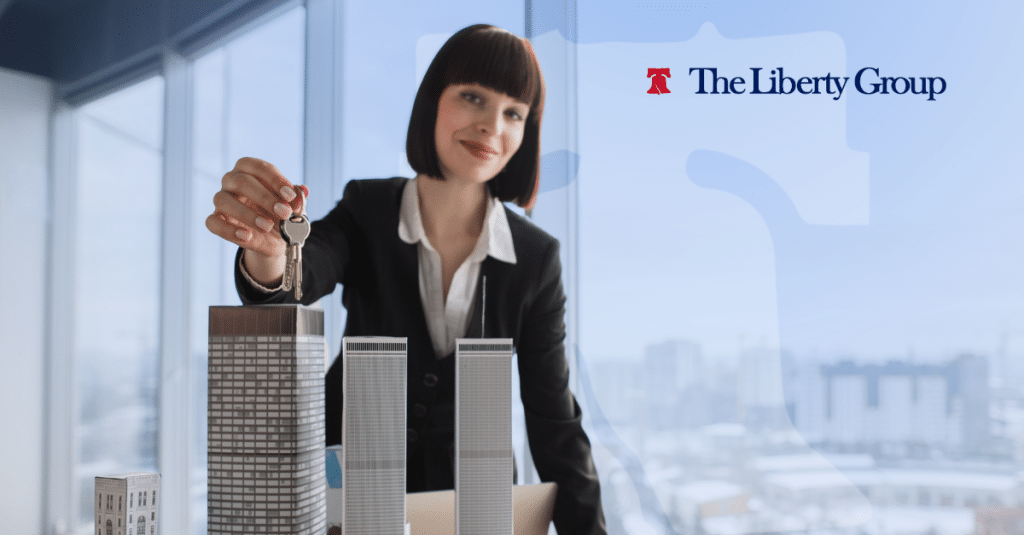 A woman in a suit holds keys over a model building in an office setting; The Liberty Group logo is in the top right corner.