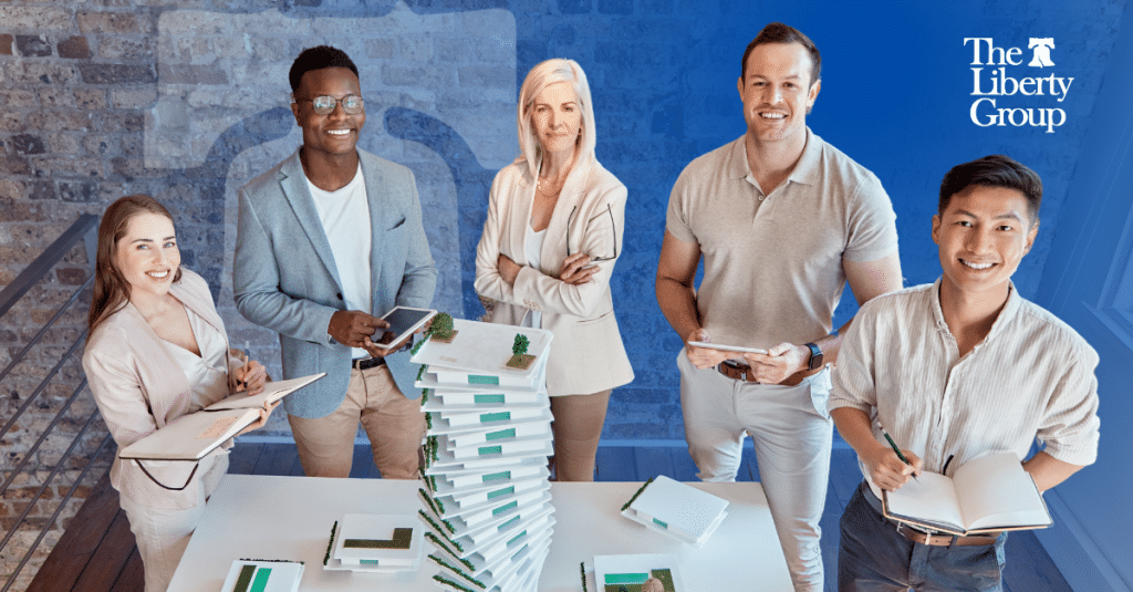 Five people standing around a table with stacked booklets, holding notebooks and tablets, with a brick wall background; The Liberty Group logo is in the top right corner.