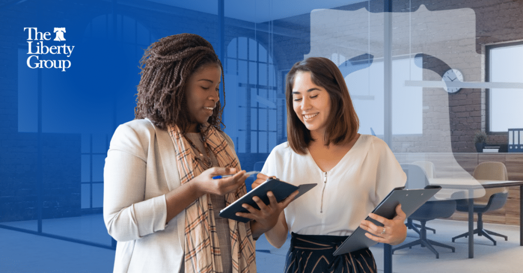 Two property management professionals discuss what's in the other's notebook. The Liberty Group logo is at the top left corner.