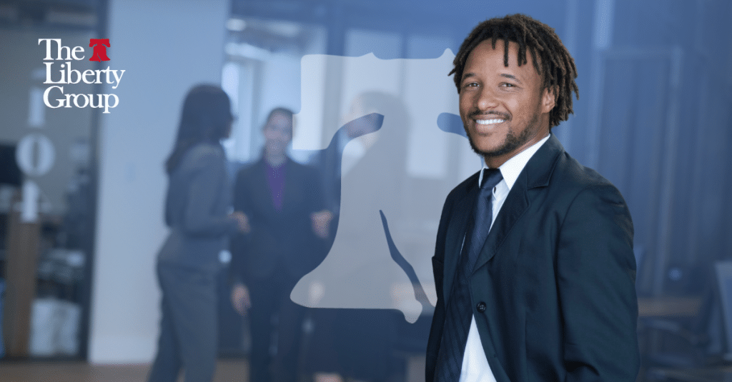 A smiling man in a suit stands confidently in an office setting, with The Liberty Group logo and blurred colleagues in the background.
