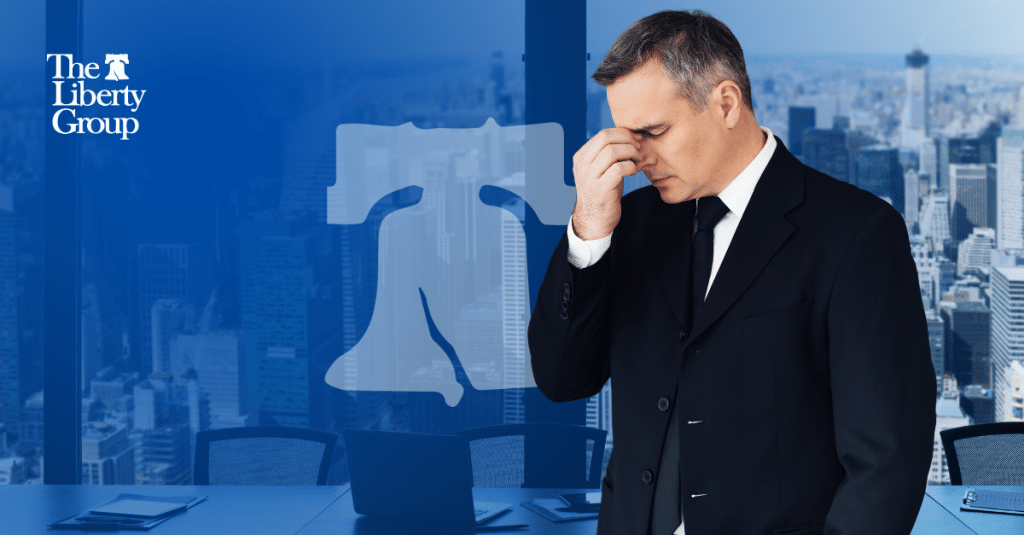 A businessman in a suit looks stressed over turnovers, standing in front of The Liberty Group logo and a city skyline.
